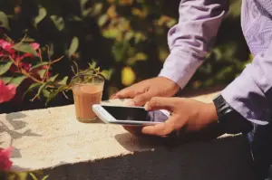 Mãos de um homem mexem no celular apoiado em uma mureta, ao lado de um copo