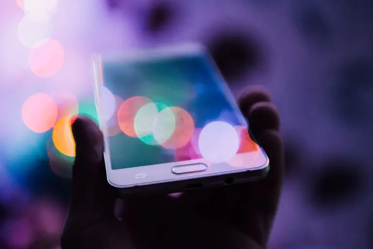 Mão segura um celular com a tela refletindo luzes coloridas desfocadas