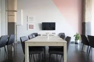 Sala de reunião vazia com mesa clara, cadeiras pretas e televisor na parede